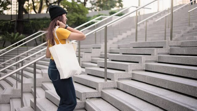 Go green Gen Z woman asia young people carrying eco tote bag reuse step walk up stairs in city life save the earth planet world care. Reduce garbage issues asian hope net zero waste the way forward.