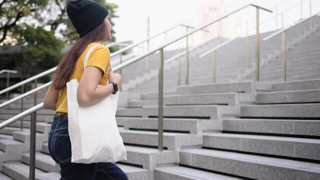 Go green Gen Z woman asia young people carrying eco tote bag reuse step walk up stairs in city life save the earth planet world care. Reduce garbage issues asian hope net zero waste the way forward.
