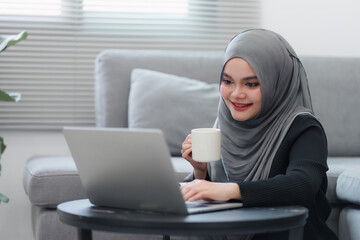 Happy Muslim woman enjoying a cup of coffee while working on her laptop at home, embodying a relaxed work from home atmosphere.