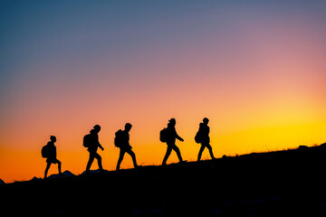 Fototapeta na wymiar Group of hikers with backpacks silhouettes walks uphill in mountains against sunset sky