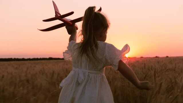 Happy little girl kid in white dress running with plane toy on wheat field at sunset sunrise horizon back view closeup. Adorable female child playing aircraft imagine pilot flying at dusk dawn meadow