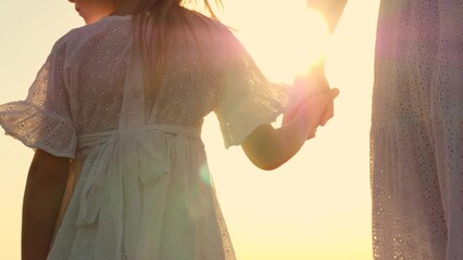 Happy family mother and daughter holding hands walking together at bright sun light sky closeup back view. Woman and girl child in elegant white dress going at natural sunlight sunny sunset sunrise