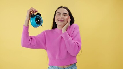 Tired female with dark hair holding alarm clock and covering mouth with hand while yawning. Charming caucasian woman looking at time and feeling like going to sleep while standing in yellow studio.