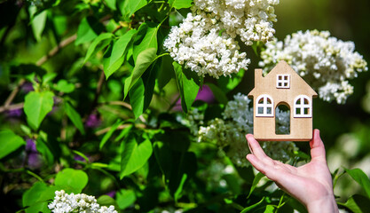 The girl holds the house symbol against the background of blossoming white lilac

