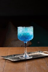 Blue color alcoholic cocktail with ice in high crystal glass on table in restaurant, dark background