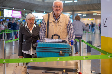 Senior travelers couple with suitcases pushing luggage trolley while walking through airport area to reach check in area and departure gate. Old senior man and woman leaving for vacation - Powered by Adobe