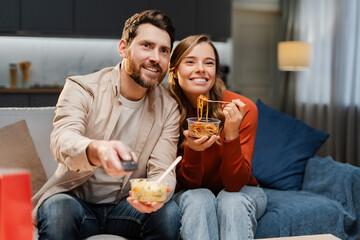 Portrait of excited couple, young man and woman sitting on comfortable sofa, holding remote control