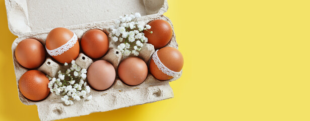 Easter eggs and white blooming flowers in carton package on yellow background, long banner. Easter...