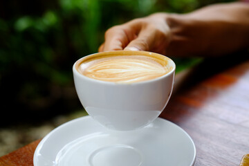 Hand holding white cup of coffee with latte in the garden.