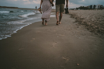 Pareja caminando por la orilla de la playa de espaldas a la cámara. Marcas de huellas en la arena de la orilla con suaves olas del mar al lado. Tienen sus chanclas en las manos.