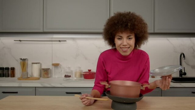 Cheerful charming African American woman with spatula smelling, enjoying aroma and taste of prepared meal from cooking pot while cooking in domestic kitchen.