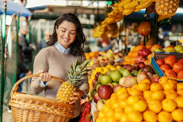 A happy woman is shopping and putting pineapple in her basket at farmers market.