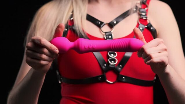Pink stylish vibrator for masturbation in the hands of a sexy girl dressed in a red bodysuit, with a leather seat belt with a chain, and fishnet stockings. Black background. Sex Shop, Adult Store
