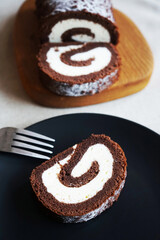 Chocolate sponge roll with vanilla cream on a wooden board next to a cut piece of rolls on a plate on a light background