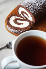 Chocolate sponge roll with vanilla cream on a wooden board next to a cup of tea