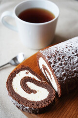 Chocolate sponge roll with vanilla cream on a wooden board next to a cup of tea