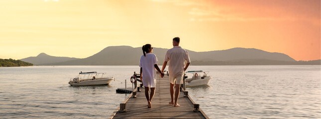 couple walking on a wooden pier in the ocean at sunset in Thailand.Caucasian men and Asian women diverse couple waking at a jetty in the ocean, man and women watching sunset together - obrazy, fototapety, plakaty
