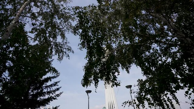 View from park on  tower in Astana, Kazakhstan. Moving camera show tower from trees.  Nur-Sultan city, Kazakhstan. People walk around the square near the Bayterek tower monument in Astana.