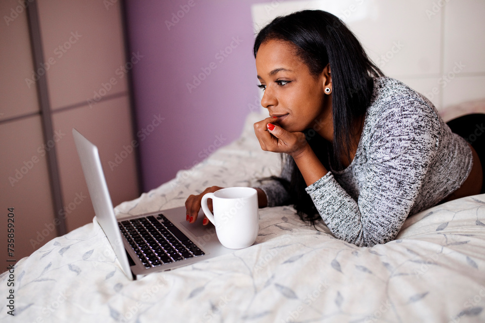 Wall mural Happy young multiracial woman works with laptop lying over bed while drinks coffee. Businesswoman, technology concept.