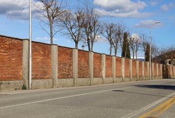 Long surrounding brick wall with  pole andconcrete columns at the road side. Bare trees, street...