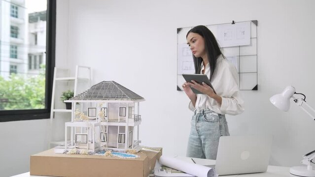 Beautiful caucasian architect focus on using tablet to check house model on meeting table with blueprint, house model and architectural document scatter around at meeting room. Immaculate.