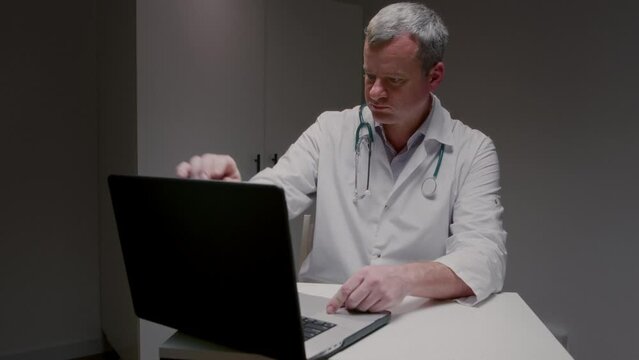 Doctor with serious expression on face opens laptop for video call with patient with health problems. Specialist sits at workplace communicating with client via online conversation slow motion