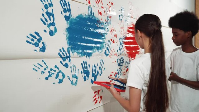 Side view of young children stamp hand with color and paint wall. high school student with mixed raced holding tray while paint room with red and blue in art lesson. Creative activity. Edification
