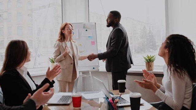 Boss shaking hand of young shy woman congratulating successful employee with promotion, hiring intern, appreciating for good work result, rewarding while business team applauding supporting colleague