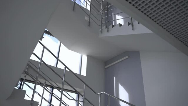 Empty staircase with shiny metal railings in hall with white walls and large windows. Minimalist interior design of multistory building