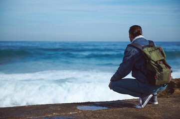 Young male traveler with backpack, admiring the dangerous splashing waves from rocky cliff. People...