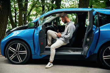 With smartphone in hands. Beautiful young man is sitting in the blue car outdoors