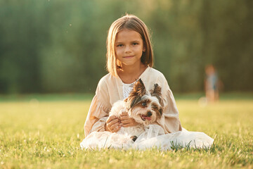 Front view, sitting together. Cute little girl is on the field with dog