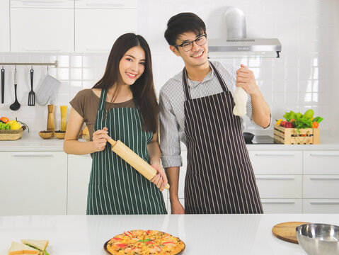 Asian man wearing eye glasses preparing pizza in modern kitchen with beautiful woman with long black hair .Asian young couple showing the dough that they made for pizza.