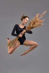young talented beautiful dancer in black bodysuit posing with bun of flowers, plants in studio,...