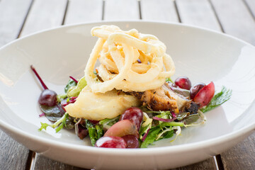 Mesclun salad with crispy squid, cod, marinated bacon, grapes and a smoked garlic dressing on white plate in restaurant