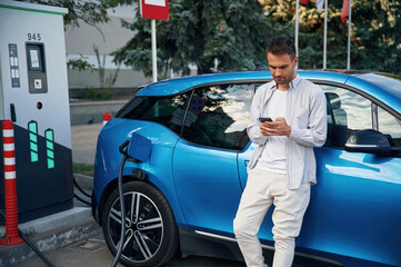 Standing with smartphone. Man with blue electric car on the charge station