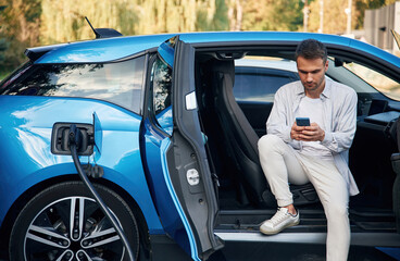 Handsome person with smartphone. Man with blue electric car on the charge station