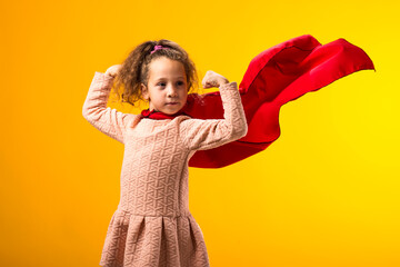 Kid girl superhero in a red cloak showing strenght gesture on yellow background. Concept of victory...