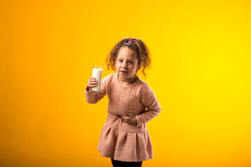 Kid girl holding glass of milk and feeling abdominal pain on yellow background. Lactose intolerance concept