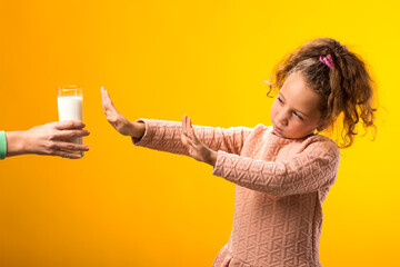 Kid girl with dairy allergy giving up of glass of milk on yellow background. Lactose intolerance...