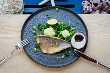 Top view of Fried Sea Bass with mixed green vegetables and mash and sauce on a dark plate with knife and fork in restaurant