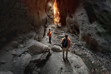 Couple of young hikers with backpacks are standing in big cave or canyon and looks at exit