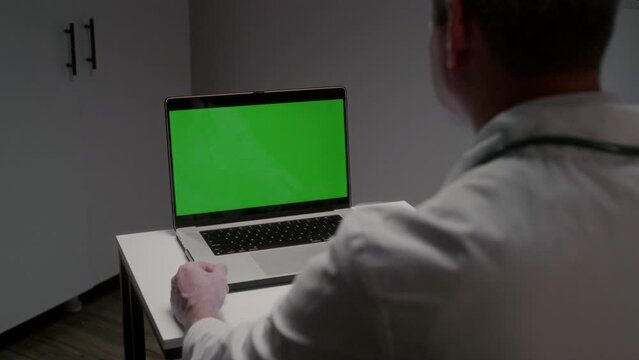 Male doctor sits at table in clinic office looking at laptop with green screen. Important profession in medical field. Work process and filling database using modern technology slow motion