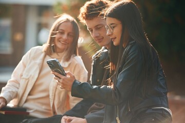 Girl is showing something in smartphone. Three young students are outside the university outdoors