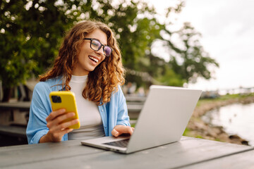 Young woman  with a laptop and phone at a table in an outdoor cafe near a lake. Education online....