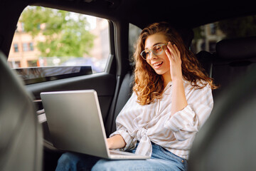 Young woman with laptop while sitting in the back seat of a car. Education online. Remote work. Business, blogging, freelance concept.