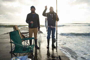 Happy, portrait and men fishing at ocean with pride for tuna catch on pier at sunset. Fisherman, friends and smile holding fish and rod in hand on holiday, adventure or vacation in nature at sea