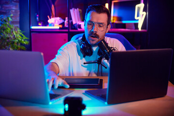 Man in vibrant studio setting speaking into microphone and typing on laptop keyboard in neon light...