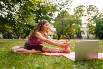 Sporty young woman practicing yoga in front of laptop, watching online tutorials in the park.  Training workout. Healthy lifestyle, sport. Online training, course.