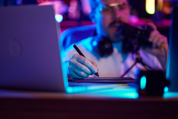 Content creator in glasses recording audio in brightly neon light, colorful home office. Selective...